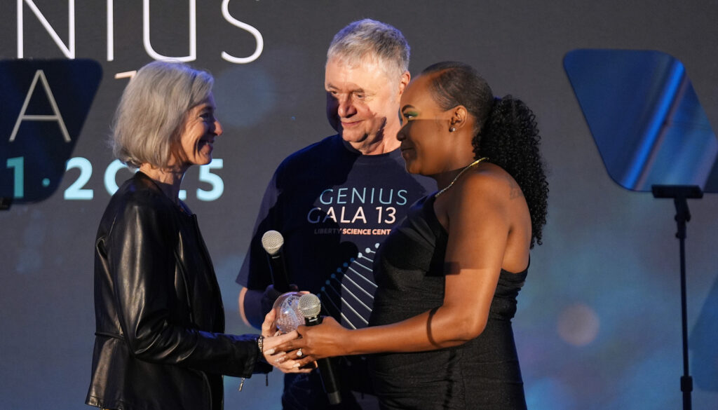Victoria Gray presents the Genius Award to biochemist Jennifer Doudna on stage. Liberty Science Center President Paul Hoffman stands between them, smiling as Doudna accepts the award.