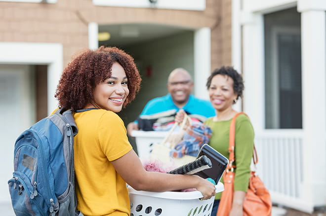 A teenage girl smiling and leaving home for college with dorm supplies.
