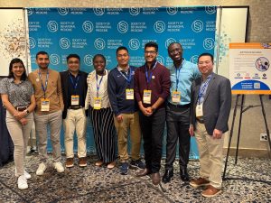 A group of researchers wearing lanyards and business clothing poses for a photo in front of a background that says "Society of Behavioral Medicine"