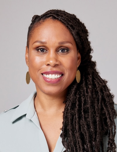 A Black woman with long dark braids and wearing gold earrings and a grey blouse smiles for a portrait against a grey backdrop.