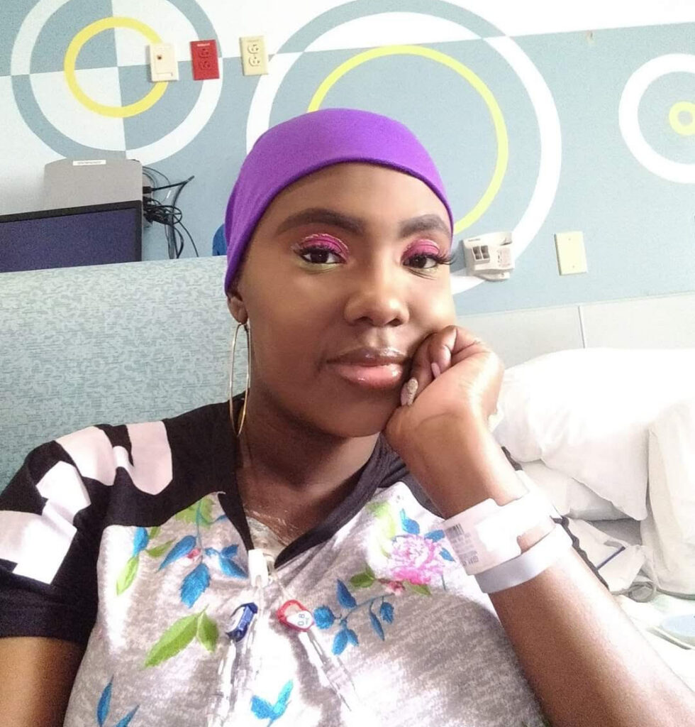 Victoria Gray wearing a purple headwrap resting her chin on her hand in a hospital bed with a medical ID bracelet and catheter visible.