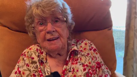 The picture shows a person sitting in a high-backed tan armchair inside what appears to be a living room or sitting room. The person is wearing glasses and a light-coloured floral shirt with red flowers.