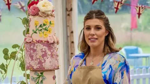 Love Productions Georgie Grasso stood in the Bake Off tent. She is wearing a beige apron and underneath it has a blue, pink and white marbled top. She is looking at the camera with her brown and blonde hair half tied up and half down. There is a cake to the left of her, which is a three tiered cake, in different shades of pink with flowers adorned on it.