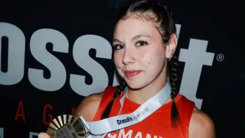 @mayhemathlete Keira McManus, a girl with dark hair tied in two plaits and wearing a red sports top, smiles as she holds up a large gold medal, which is around her neck.