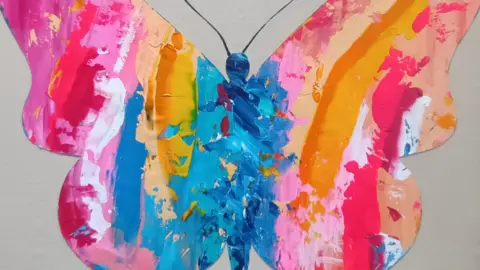 Allegra Dams A butterfly-shaped artwork called My Angel by Allegra Dams is painted in bright pink, orange, yellow and blue colours against a plain background.