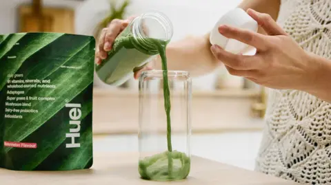 Huel A green nutritional drink is being poured into a glass. The setting appears to be a bright, modern kitchen or dining area with a blurred background.