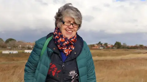 Family handout A woman standing in a field on a cloudy day. She has short grey hair and is wearing glasses along with a green and black coat with a blue scarf with red foxes on it.