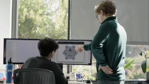 Stanford University This image shows two people working at a desk in a bright office or research environment with large windows and natural light. One person is seated in front of a dual-monitor workstation, while another stands nearby pointing toward a detailed molecular or protein structure displayed on one of the screens. The monitors show scientific visualization software, with one screen displaying a complex three-dimensional biological model and the other containing text or data. Various desk items, including a water bottle, mug, computer peripherals, and potted plants, are visible around the workspace.
