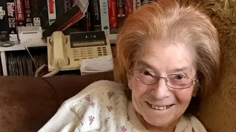 Family photo A elderly woman smiling at the camera. She has thin black frame glasses and short blonde/light brown hair. She is wearing a white top with small flowers on them. There is a landline telephone behind her and she is sitting on the sofa