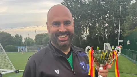 Supplied A man smiles straight at the camera. He is standing on a football pitch and holding a trophy, while wearing a black jumper over a green football top.