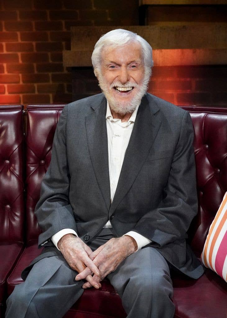 Dick Van Dyke sits on a red leather sofa in a grey suit with a white shirt and white beard on set of a CBS Original dedicated to his career.