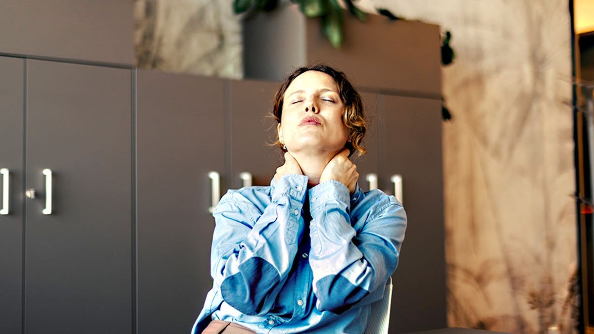 Businesswoman massages her neck with eyes closed in modern office.