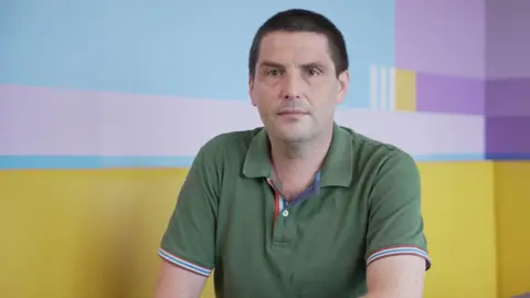 Dr Paul Evans is sitting at a table in front of a yellow seating area looks into the camera. He has dark hair and brown eyes. He's wearing a green polo shirt with blue and red piping around the sleeves.