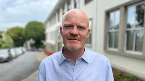BBC Deputy Gavin St Pier - A bald man with a ginger and grey speckled beard. He's wearing a blue buttoned shirt with the collar undone.