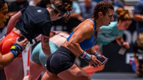 @mayhemathlete Keira - a girl with brown hair, blue sports top and black shorts - leans forward as if about to start running in a race as other competitors line up next to her.