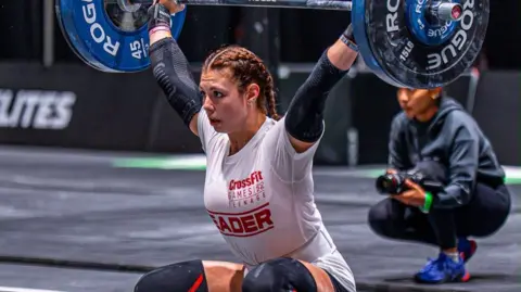 @mayhemathlete Keira McManus - a girl with dark hair tied in two plaits, wearing a white sports shirt, shorts and knee pads - crouches down while lifting a barbell with large round weights on either end.