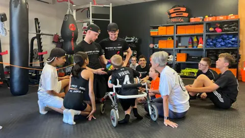 A group of children with  Bobby and Shay looking at a multicoloured wheel. They are in the gym with punchbags in the background and apparatus. There is a woman wearing a white t-shit which is Bobby's nan.