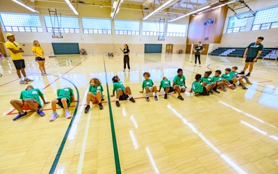 A row of children in green shirts sit spaced apart across a large wooden gym floor, stretching or participating in an activity while several camp counselors supervise.