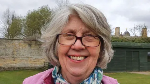 Family handout A woman standing outside on a grey day. She is smiling at the camera. She has short grey hair and is wearing glasses along with a pink jumper and a blue and green scarf.