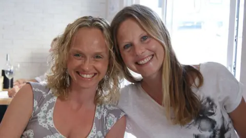 Family photo Two women sit together and smile at the camera. Their heads are leaning together and they are smiling. They are in a restaurant and behind them is a table with a wine bottle and wine glass on it.
