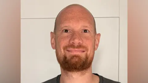 Gary Prince A man looks straight at the camera while smiling. He is standing in front of a white door and wearing a dark grey T-shirt.