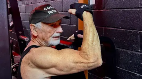BBC Mick Wadge flexes his right arm while using a weight-training machine at the gym.