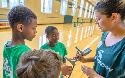 A close-up view of a female instructor in a green Kinesiology shirt holding a silver handgrip strength tester while speaking to three young boys in a gymnasium.