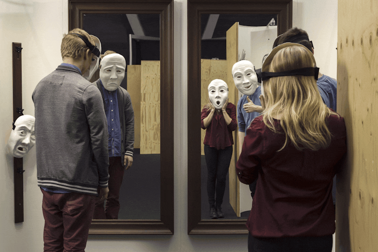 People standing looking in mirrors wearing giant masks.