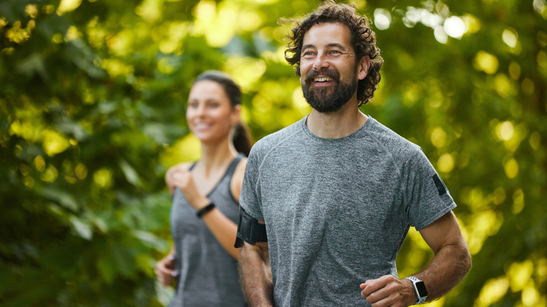 A man and a woman running outdoors under trees, with wearable devices on their wrist.