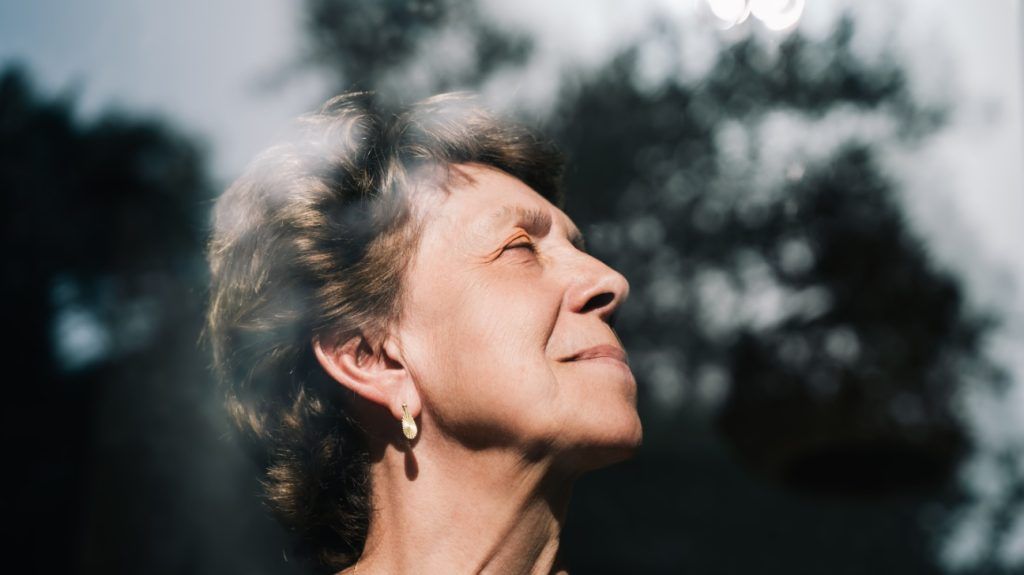 An older woman looks up and to the side as the sun shines on her face through a window