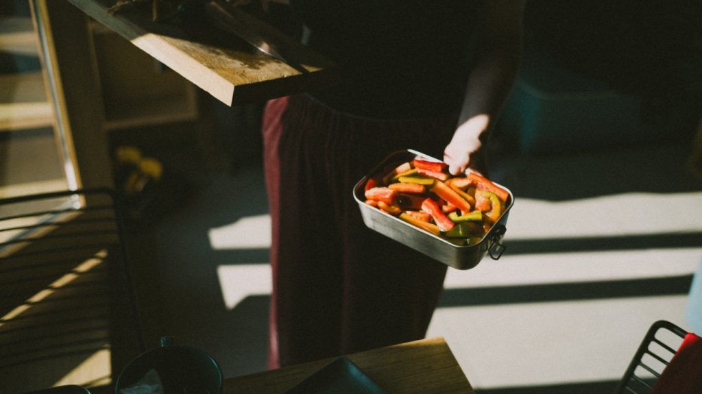 A woman holding a lunchbox of sliced bell peppers as part of a low protein lunch in the kitchen in sunlight