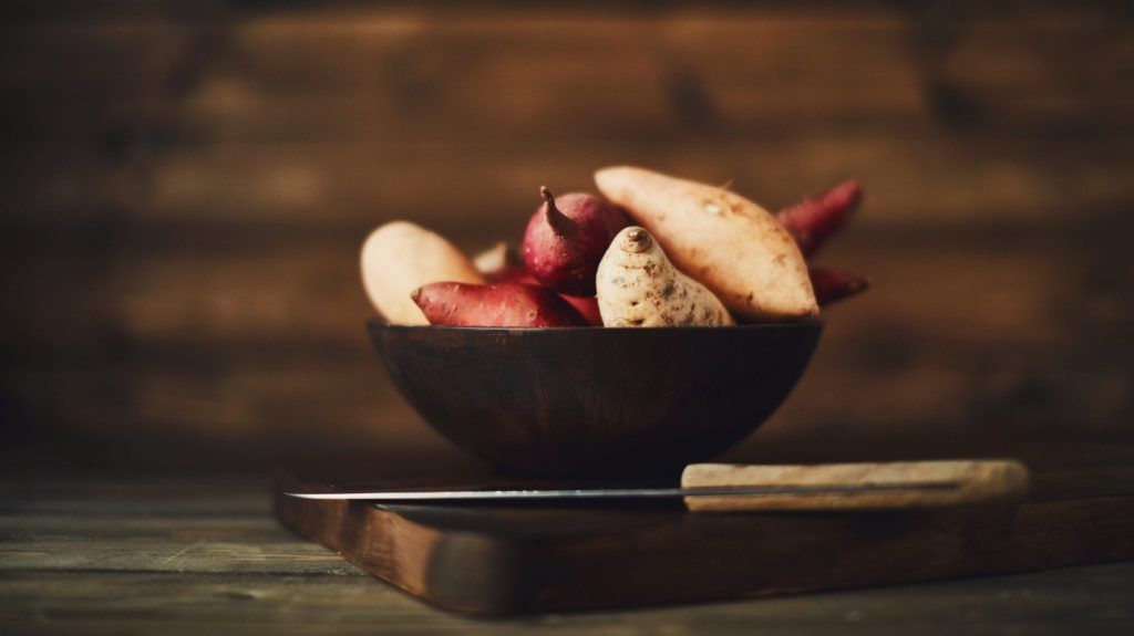 A bowl of sweet potatoes on a chopping board with a knife underneath on a rustic table