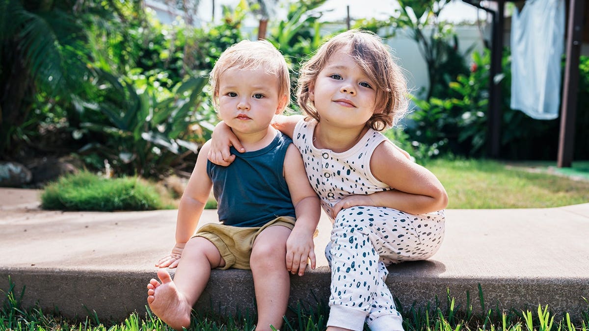 Three-year-old girl embraces her one-year-old brother on a paved walkway in a backyard garden.