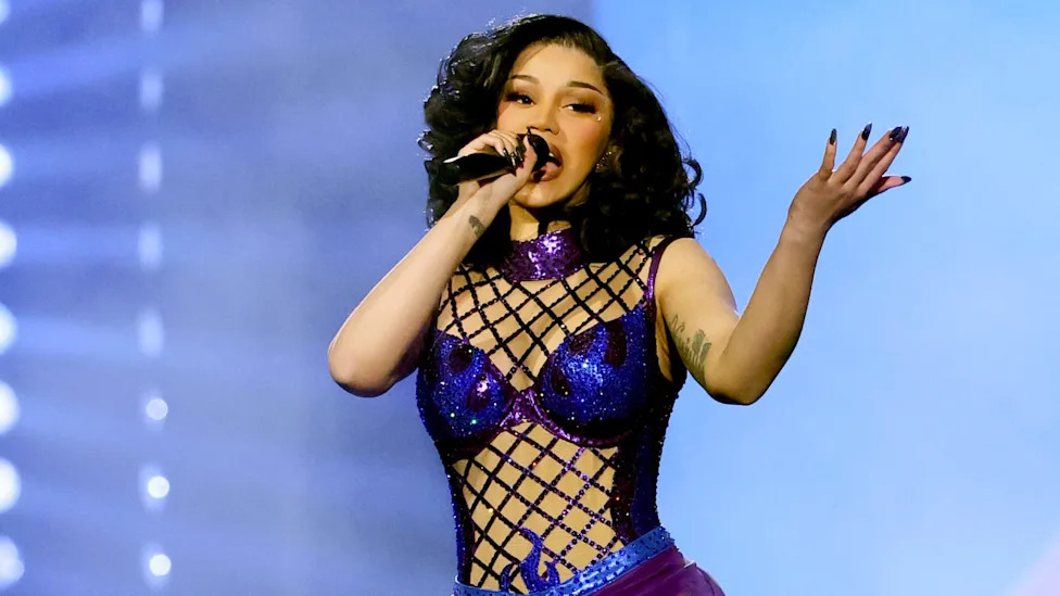 Cardi B performs in a purple, netted outfit sings on stage while holding a microphone.