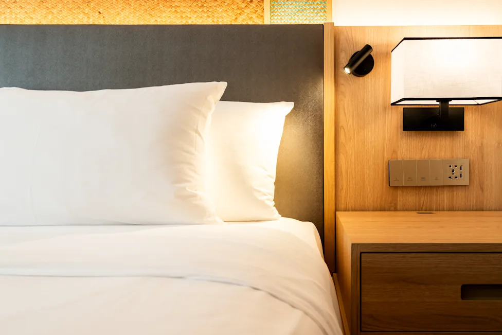 Detail of a clean and modern hotel room showing the bed with white pillows, wooden nightstand, fabric wall lamp, and a power outlet, switch panel.