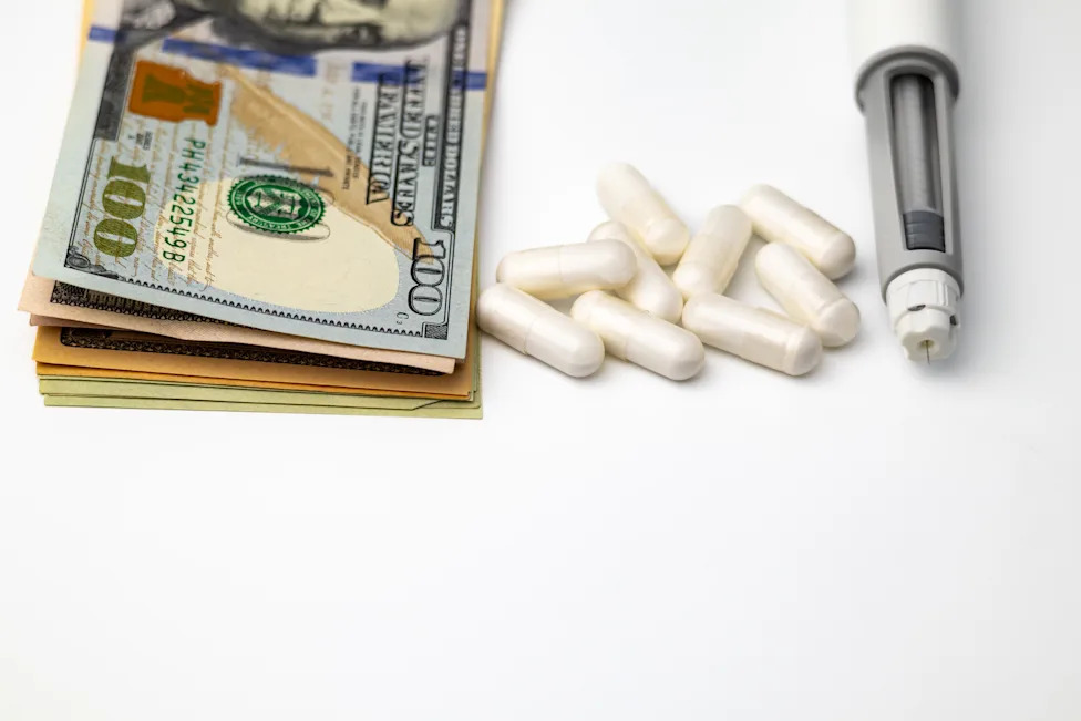 A stack of hundred-dollar bills next to white GLP-1 capsules and an injector pen, representing the cost of weight-loss medication.