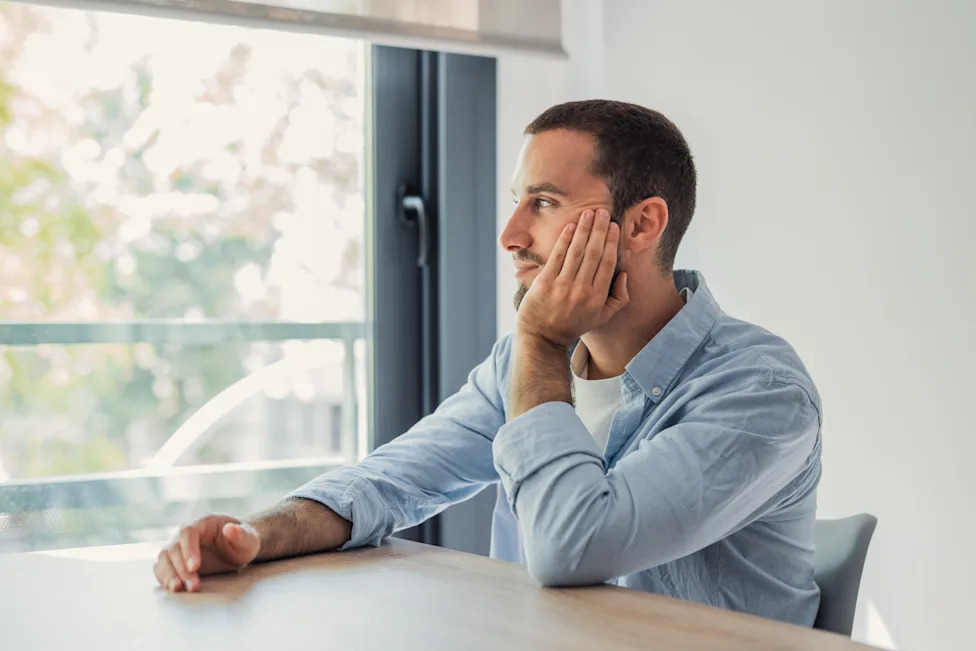 Young man sitting at wooden table, resting his head on hand, looking out a window, thinking and reflecting in a calm indoor environment