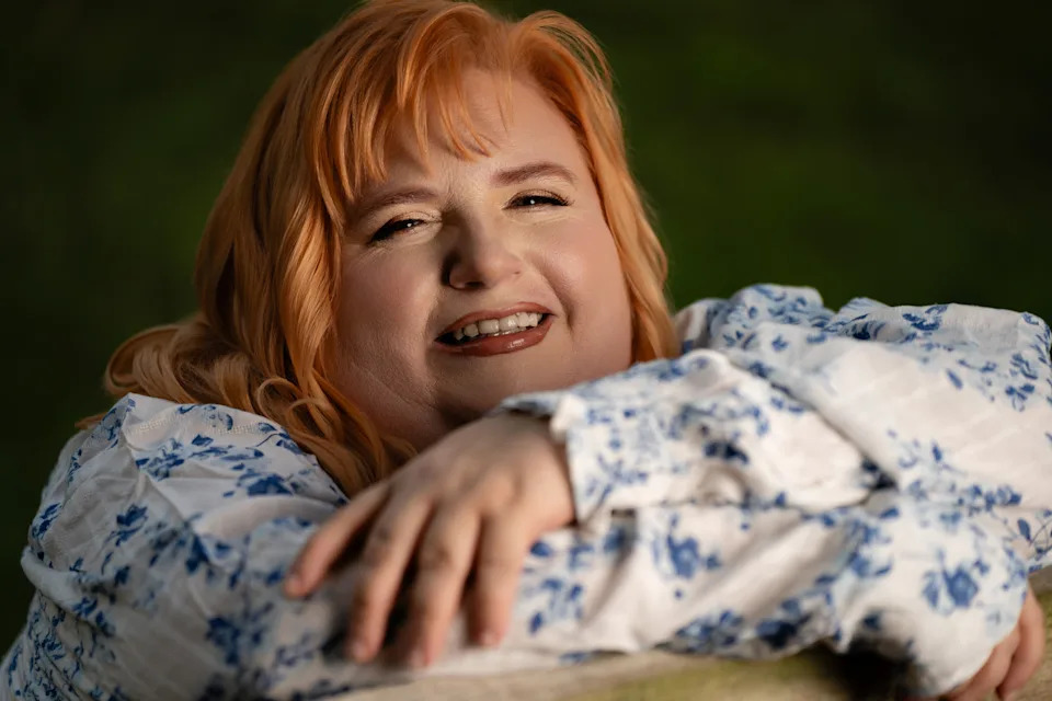 Woman with long strawberry blonde hair in a blue and white floral top looks toward the camera with a thoughtful expression against a green background.