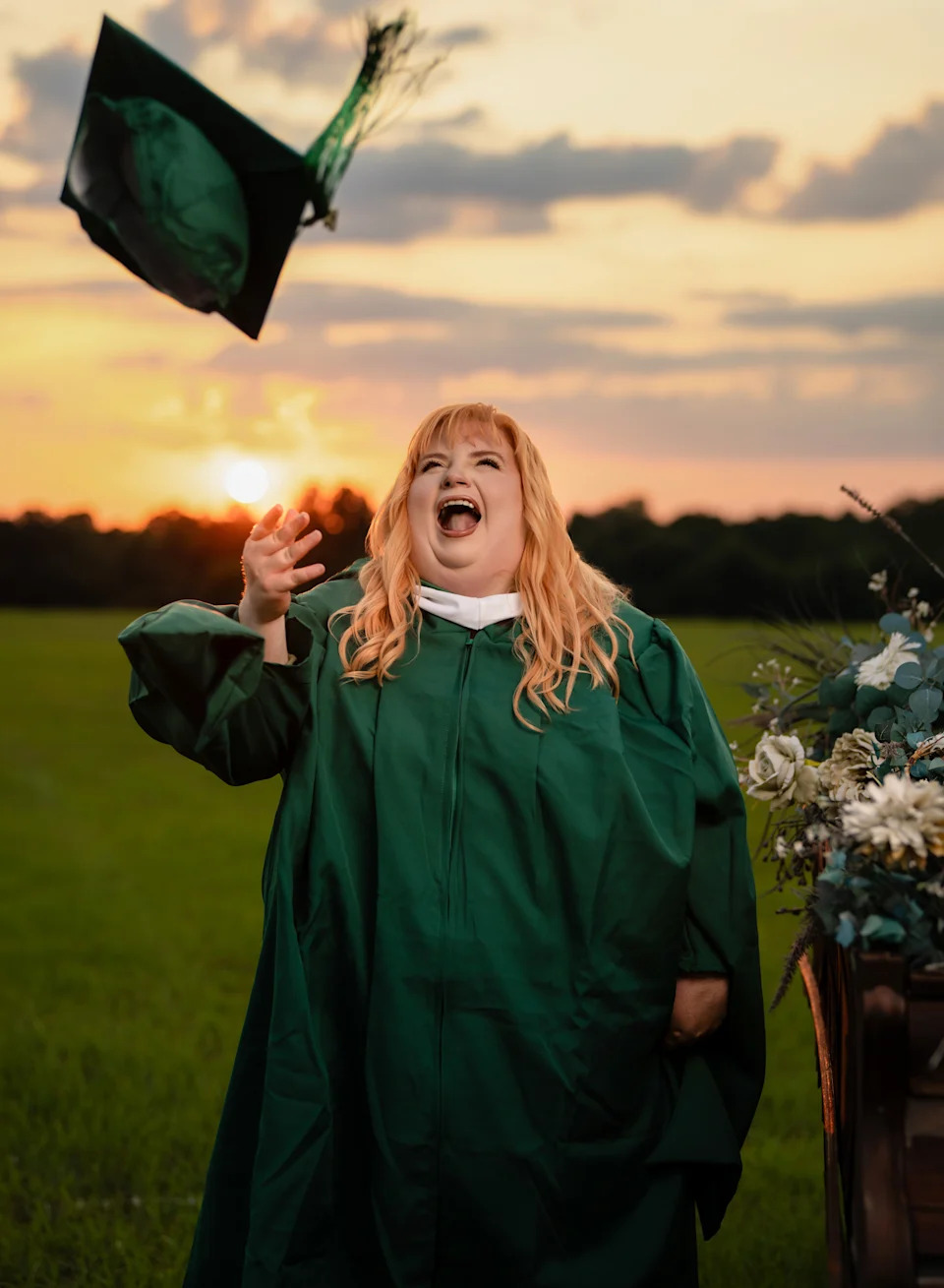 Woman in a green graduation gown throws her cap into the air and laughs against a golden sunset over an open field.
