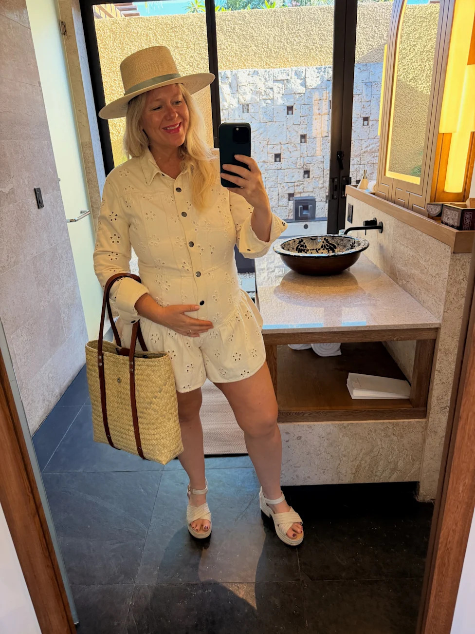Pregnant writer Brittany Anas takes a full-length mirror selfie while wearing a white eyelet romper and sun hat.