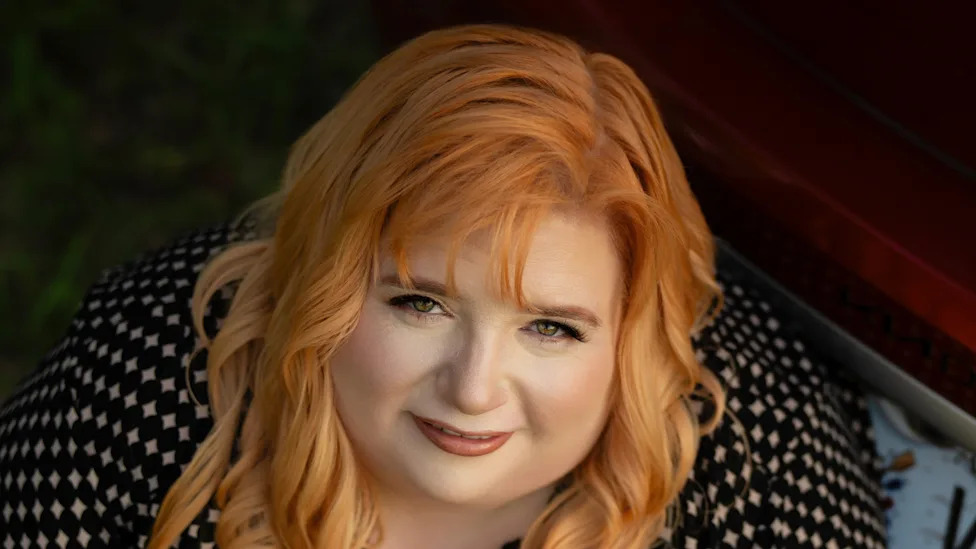 Woman with long red-gold hair in a black and white diamond-print top smiles softly at the camera in warm sunlight.