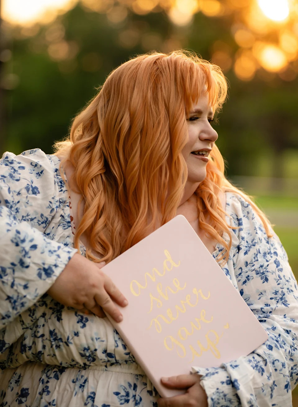 Woman with long strawberry blonde hair holds a pink notebook reading "and she never gave up" during golden hour sunlight.