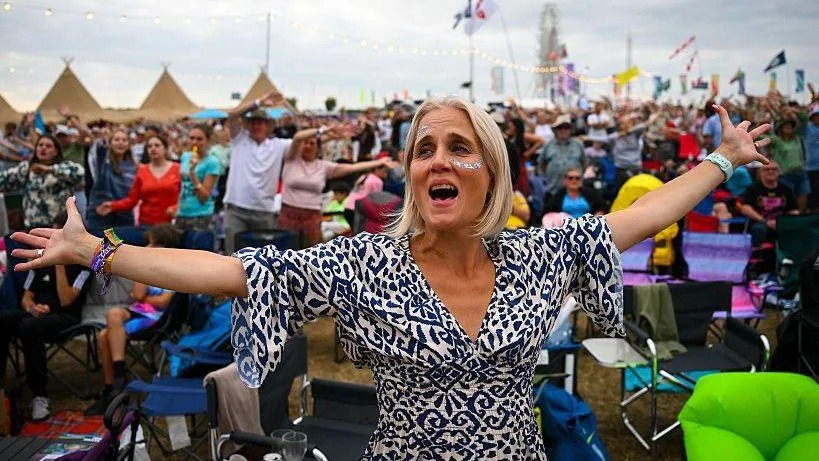 A woman with blonde hair and a blue and white dress, with her arms outstretched and singing at CarFest 2025. Behind her is a large crowd of men and women, some also swaying to music.