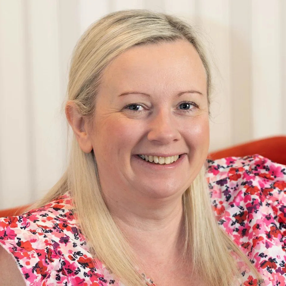 Lisa, a woman with long blonde hair in a pink and white floral top, smiles at the camera in front of a plain white background