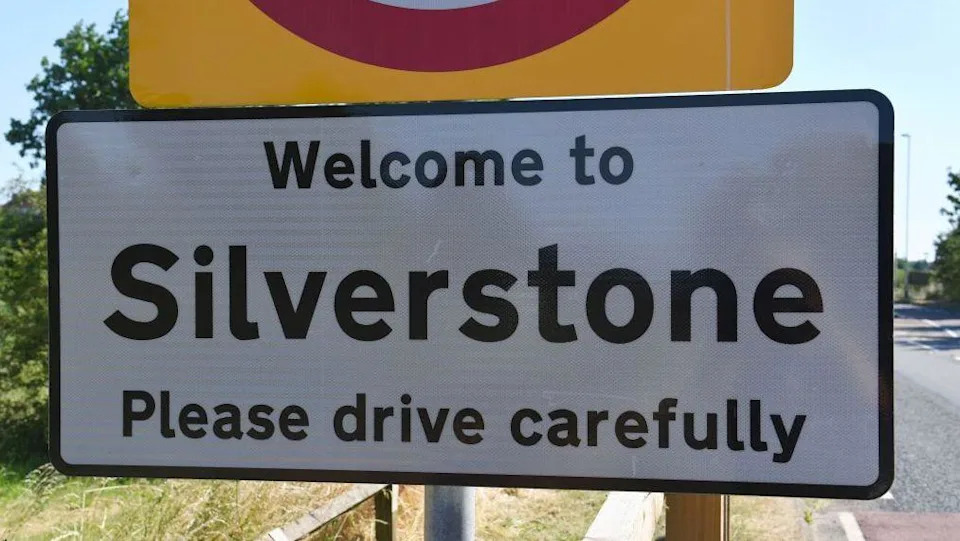 A road sign which says welcome to Silverstone please drive carefully in black letters on a white background. It is positioned on a verge to the side of a tarmacked road.