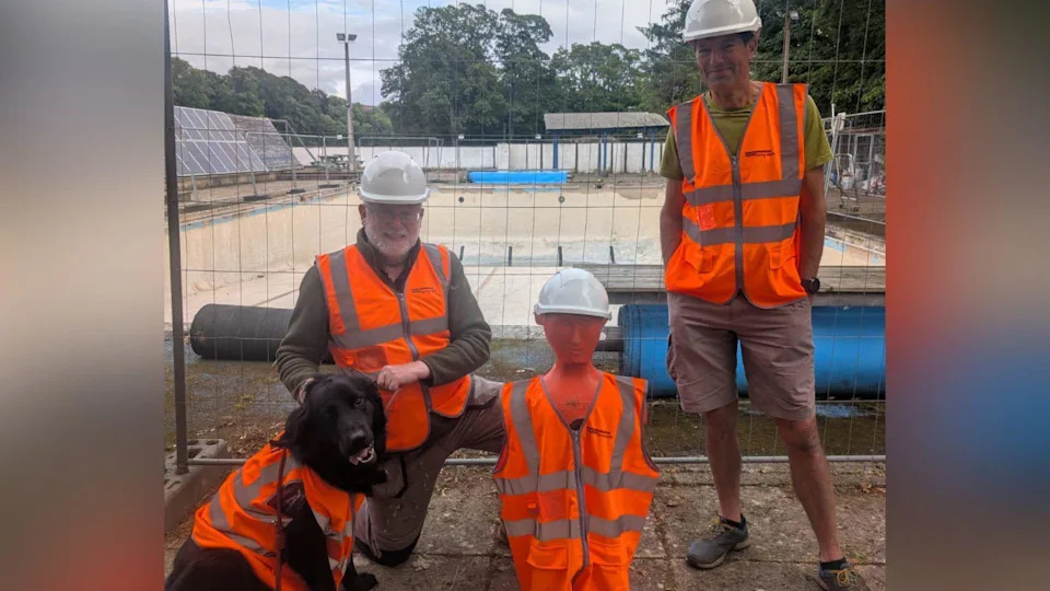 Two men, a dog, and for some reason a crash test dummy, are all donning orange high-visibility jackets. The men are wearing hard hats. The dog doesn't get one. Behind them, surrounded by construction fencing, is the open-air pool - completely dry and empty.