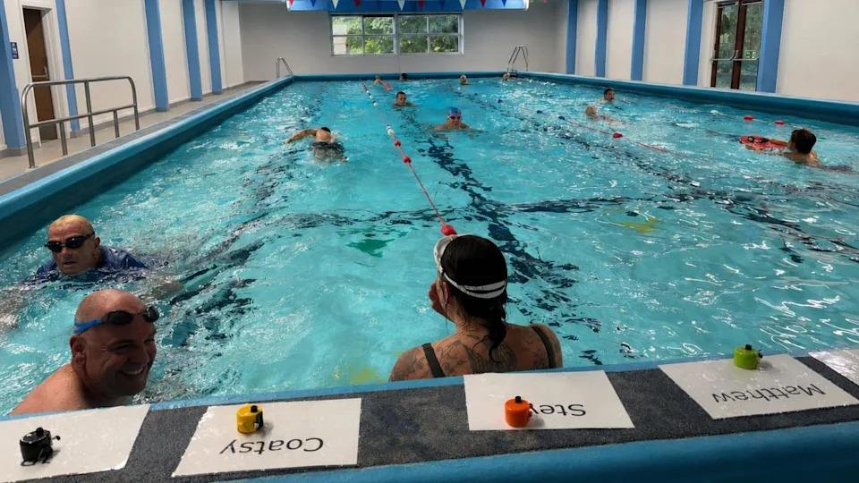 The fundraising swim when it was ongoing. In relay style, one swimmer waits in the water whilst the other swims their lengths. The water is crystal-clear, chlorinated blue, which ripples as the swimmers power through. Some wear goggles.