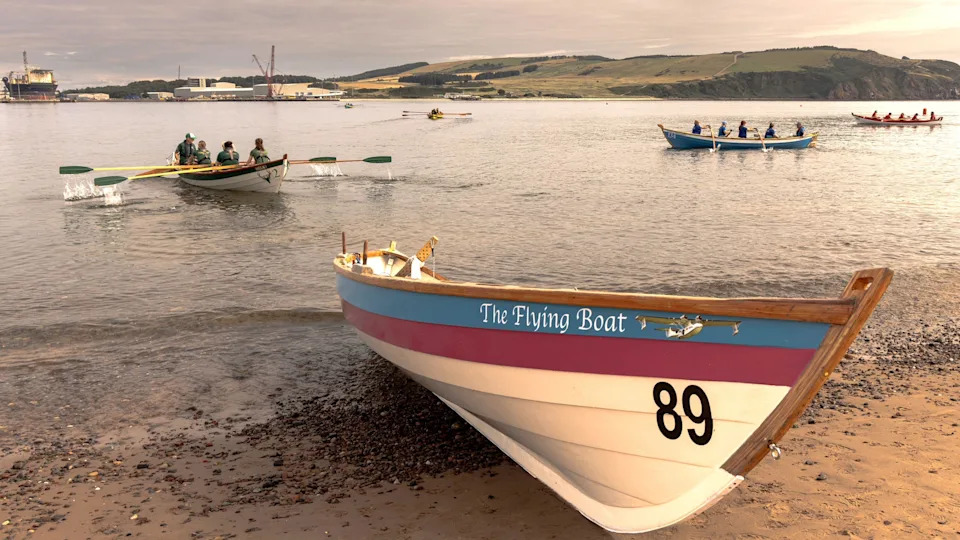 A painted skiff named The Flying Boat rests on a sandy shoreline while several crews row traditional skiffs across calm water during a regatta.
