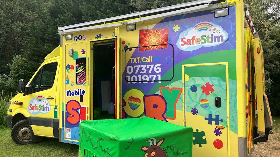 A yellow ambulance which has been been transformed into a sensory van. On the side of the van there are colour stickers and symbols. The door of the van is open and there is a green box next to it.
