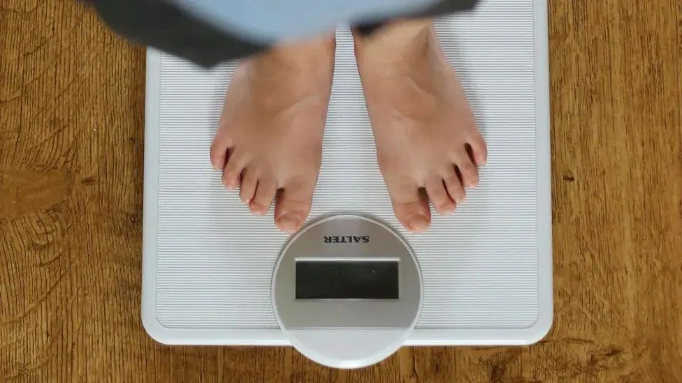 The image shows a close-up view of a person's bare feet standing on a white digital bathroom scale placed on a wooden floor.